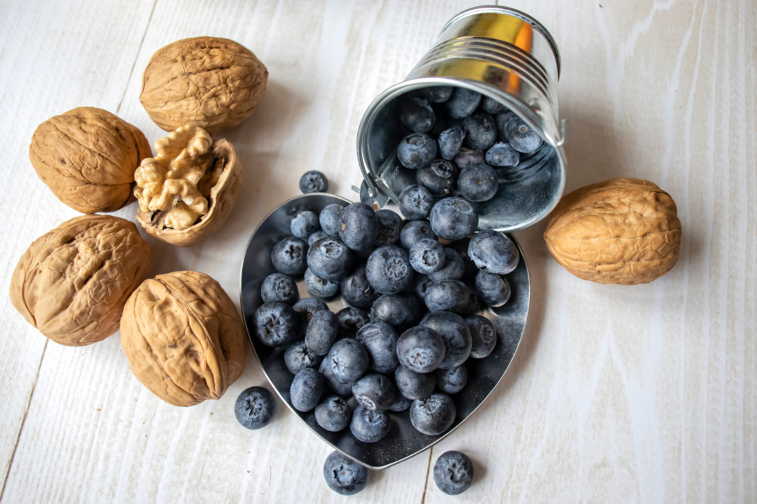 Blaubeeren in einer herzförmigen Schüssel und einem kleinen Blecheimer, daneben Walnüsse auf einer Holzplatte