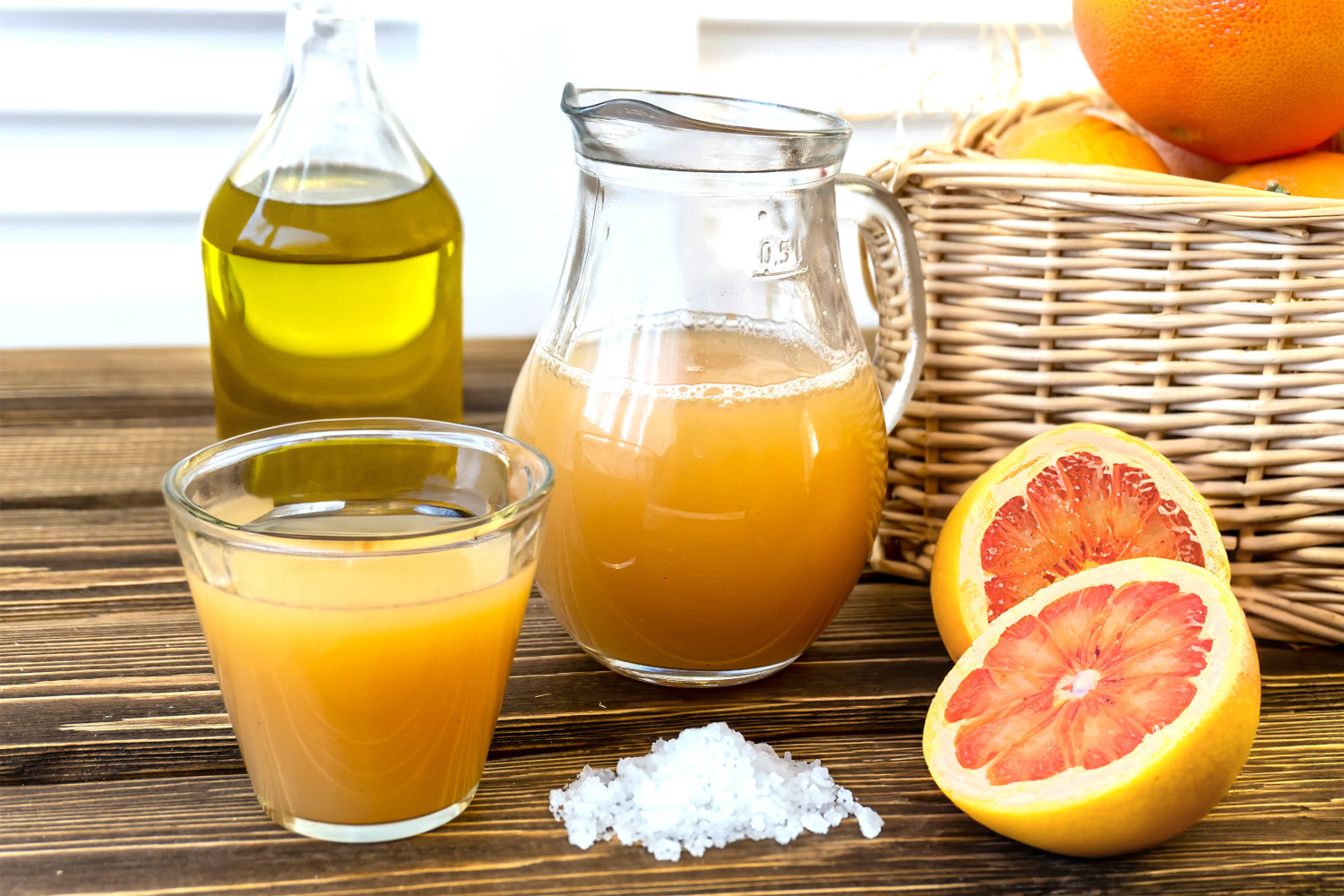 Carafe of fruit juice, beside it a filled glass, a basket of oranges in the background, coarse Epsom salts on the table, bottle of olive oil