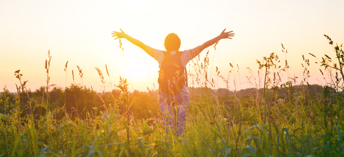 Person mit erhobenen Armen von hinten in einem Feld, Sonnenuntergang