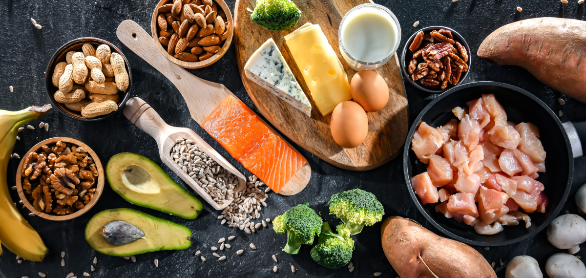 Biotin-containing foods such as salmon, poultry, avocado, dairy products, broccoli, bananas and nuts in bowls and on a slate board.
