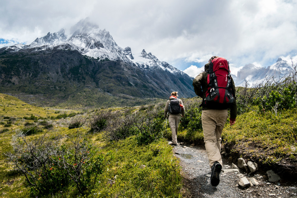 Two hikers on a stony path through green, heading towards a mountain with a white peak, rugged surroundings