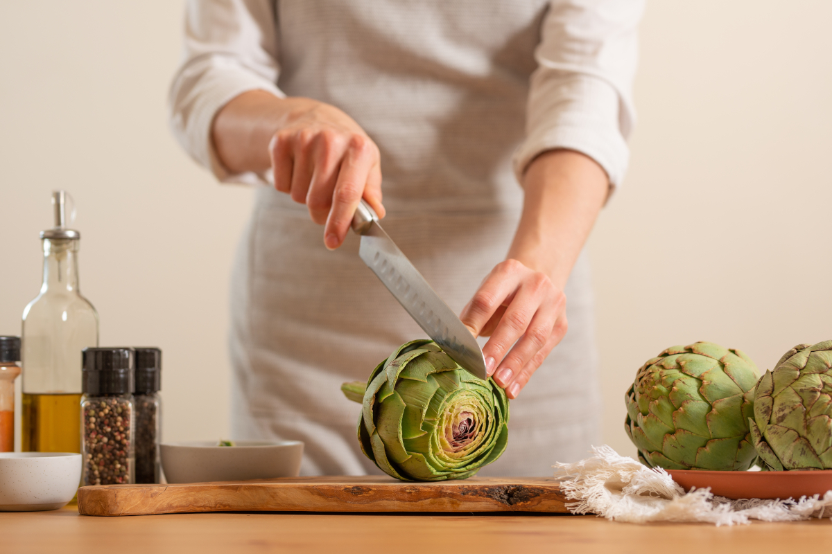 Artichoke on a chopping board being cut with a knife, beside spices and oil
