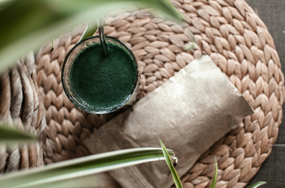 Top view of a glass with frothy Spirulina shake on a round woven mat, beside a lying doypack, narrow leaves blurred in the foreground