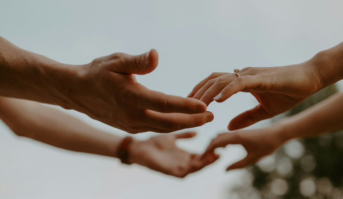 Foto: Zwei Paar Hände, bei denen sich nur die Fingerspitzen berühren. Eine Hand trägt einen schmalen Ring mit Edelstein