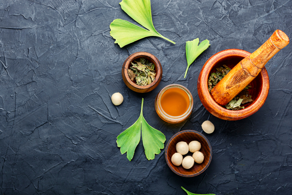 Illustration: Top view of small dark wooden bowls containing ginkgo in various forms, including dried herb in a mortar; ginkgo leaves used as decoration