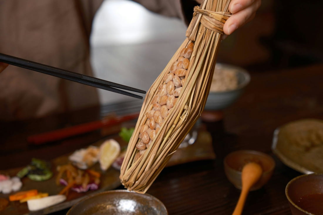 Natto in a straw casing being held over a bowl. Two chopsticks are in motion, pushing the natto into the bowl. Various vegetables, cut into small pieces, are visible in the background