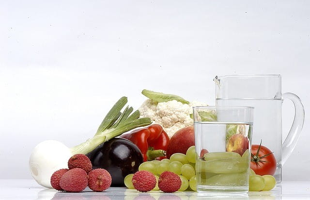 Photo of fruit and vegetables such as lychees, grapes, tomatoes, peppers, fennel, aubergine and cauliflower next to a carafe and a filled glass of water