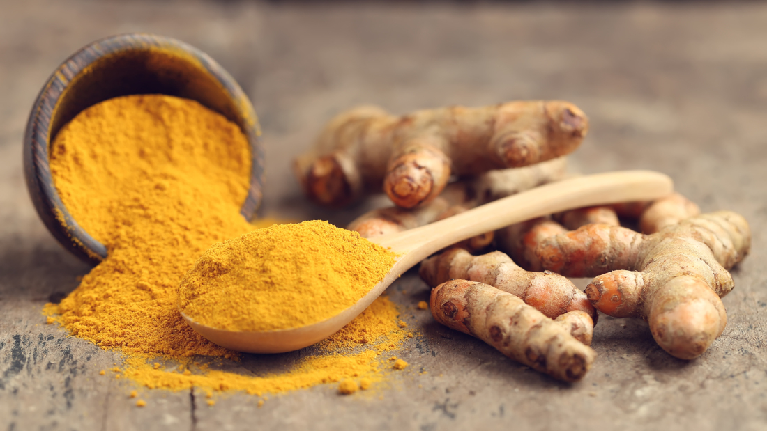 Photo of ground turmeric in a bowl with a spoon, next to a whole root, on a wooden board