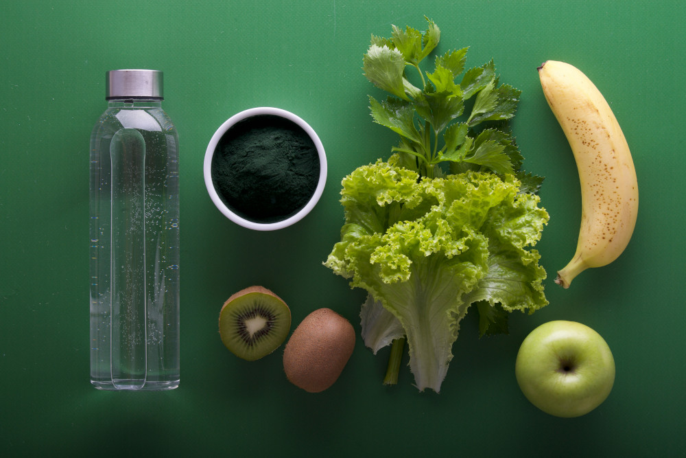 Bouteille d'eau, poudre verte dans un bol, kiwi et banane coupés, pomme verte et herbes sur un fond vert, vue de dessus.